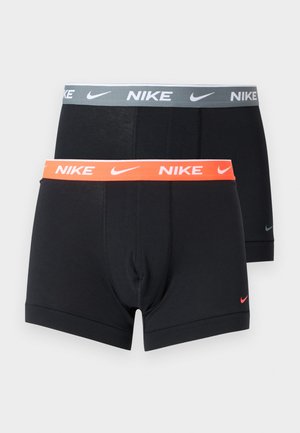 Homme grand debout pieds nus sur un sol recouvert de moquette, portant des boxers noirs Nike, regardant vers sa gauche, les mains dans le dos.