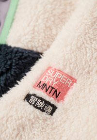 Veste en polaire avec un mélange de panneaux rose clair, bleu marine et vert. Inclus des étiquettes brodées en rose et noir avec un texte de marque.