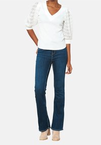 Biała bluzka z koronkowymi bufkami, dekoltem w kształcie litery V, w połączeniu z ciemnoniebieskimi dzwonami. Modelka nosi jasnobeżowe buty na słupku.