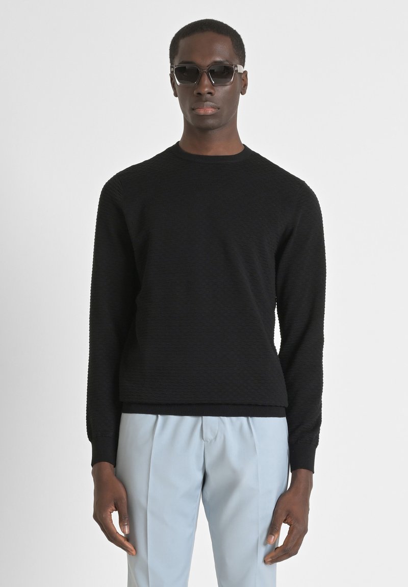 Maglione nero a trama con colletto rotondo e maniche lunghe, abbinato a pantaloni azzurri chiari. Modella che indossa occhiali da sole scuri.