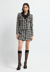 Vestido de blazer a cuadros en blanco y negro con cuello, frente abotonado y mangas largas. Combinado con calcetas negras hasta la rodilla y zapatos de tacón alto.