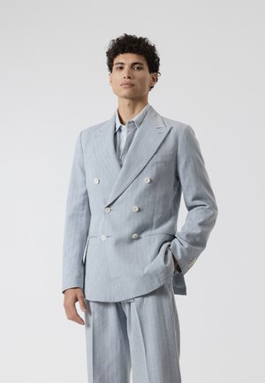 Joven con cabello rizado que lleva un traje cruzado gris claro y camisa azul clara, de pie con una mano en el bolsillo.