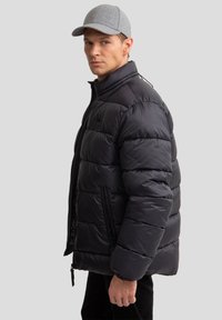 Melnā pūkaina jaka ar augstu apkakli, kas aprīkota ar horizontāliem quilted posmiem un sānu rāvējslēdzēju kabatām. Modelis valkā pelēku cepuri.