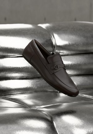 Mocassin en cuir marron foncé avec une boucle décorative, placé en angle sur une surface argentée brillante capitonnée.