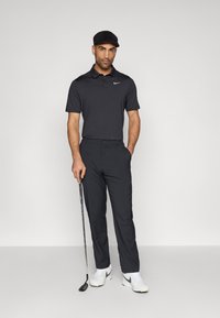 Polo shirt gris foncé à manches courtes, associé à un pantalon gris foncé. Le joueur tient un club de golf, portant des chaussures blanches Nike et une casquette.