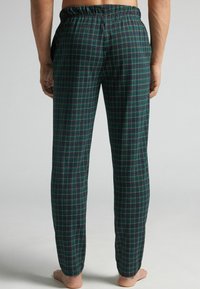 Hombre con pantalones de pijama de cintura elástica en cuadros verde y azul marino, de pie descalzo con las manos relajadas a los lados, visto desde atrás.