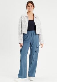 Gekürzter Tweed-Jacke in Weiß und Grau über einem schwarzen Oberteil, kombiniert mit blauen Cargo-Hosen mit Seitentaschen und einer lässigen Passform.