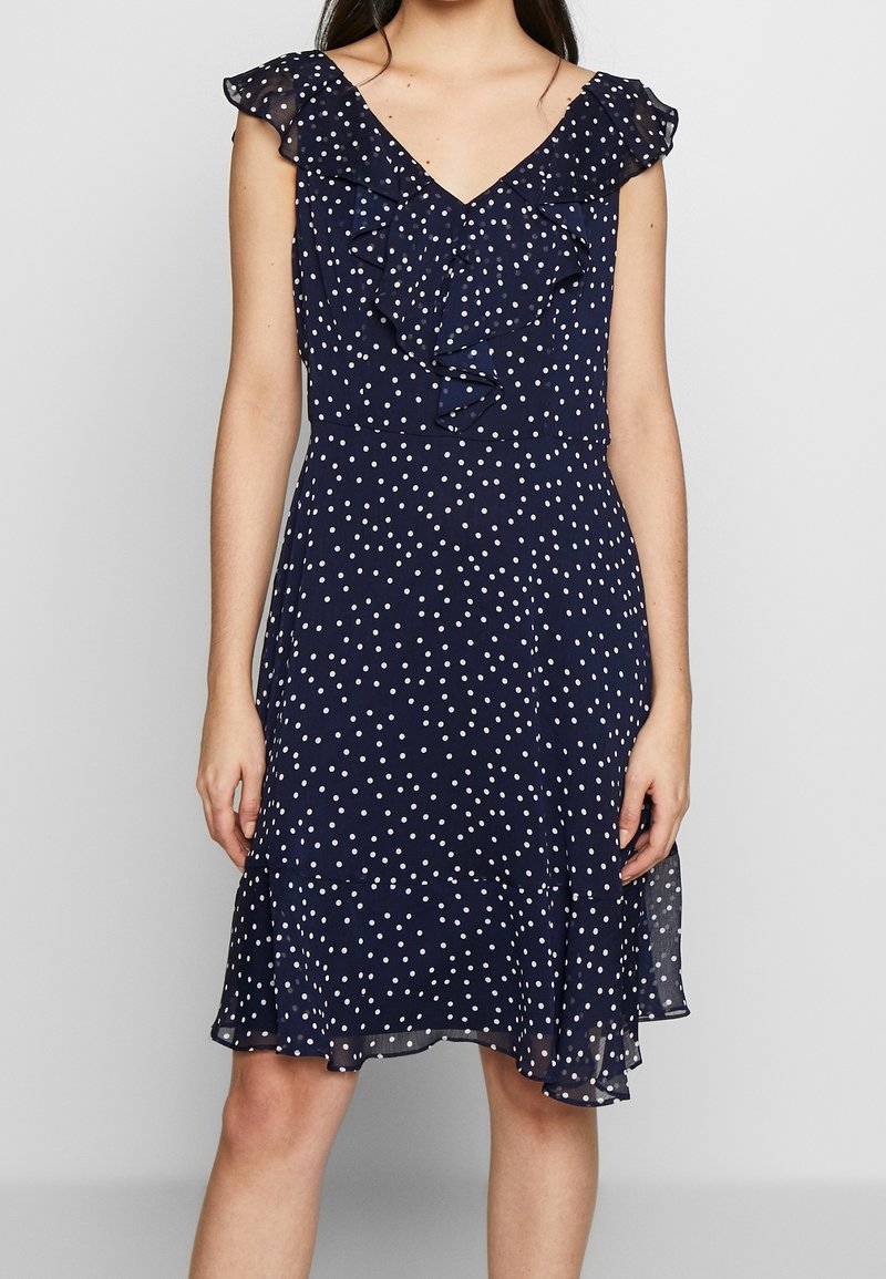 Navy dress with white polka dots, V-neck, ruffled cap sleeves, and a flared hem. Made of lightweight fabric with a semi-sheer finish.
