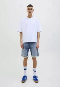 Jeune homme debout portant un t-shirt blanc surdimensionné, un short en denim roulé, des chaussettes rayées et des baskets bleues, sur fond uni.