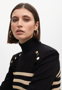 Crni kader s dugim rukavima s zlatnim horizontalnim prugama, s dodatnim gumbima na ramenima. Glatka tekstura i prianjajući kroj.