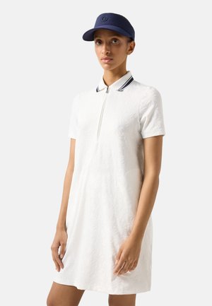 Kobieta w białej sukience polo z krótkim rękawem o delikatnej kwiatowej fakturze i granatową czapką, stojąca na jednolitym jasnym tle.