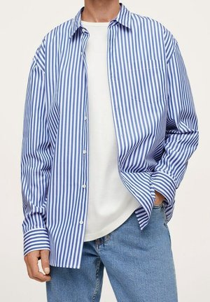 Man wearing open blue and white vertically striped button-up shirt over a plain white t-shirt with blue jeans, hand in pocket.