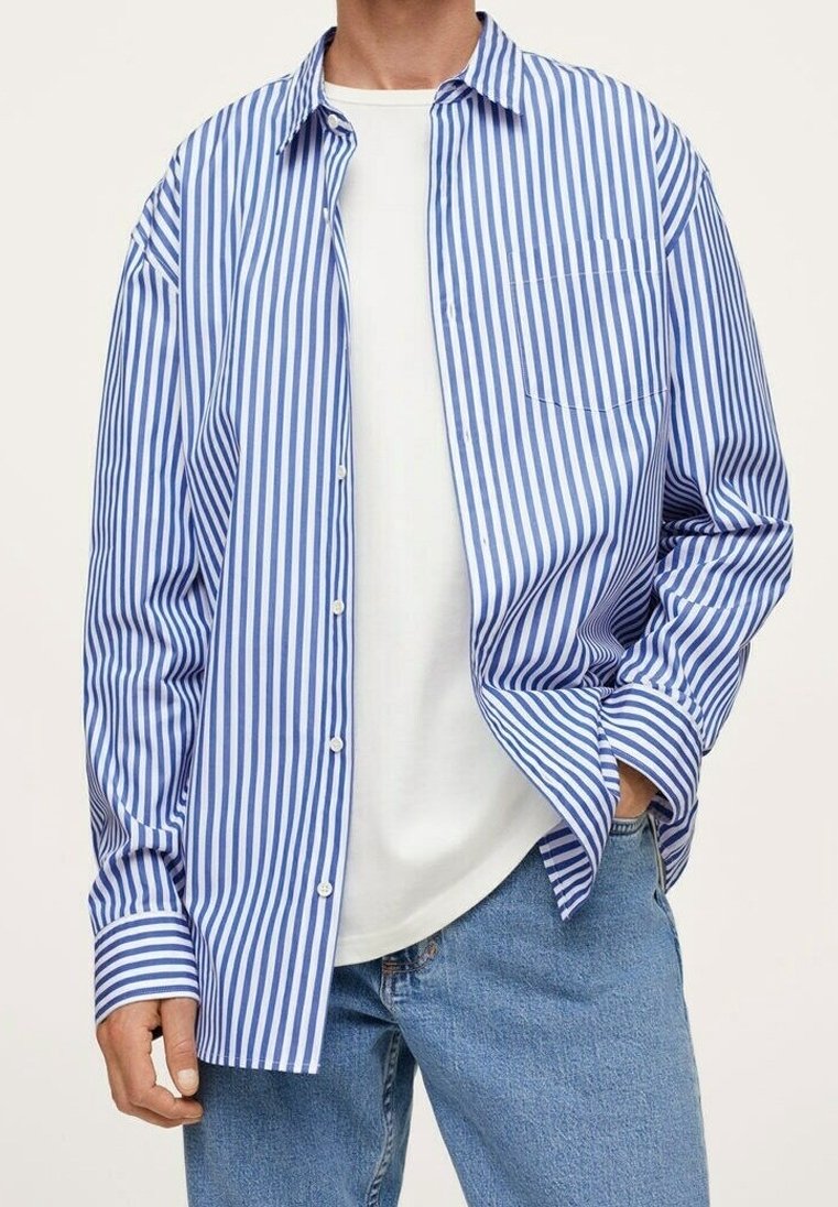 Homme portant une chemise bleue et blanche à rayures verticales ouverte par-dessus un t-shirt blanc uni avec un jean bleu, main dans la poche.