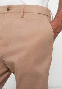 Beige skreddersydde bukser laget av glatt stoff med knappelukking, med en sidelomme og subtile søm detaljer.