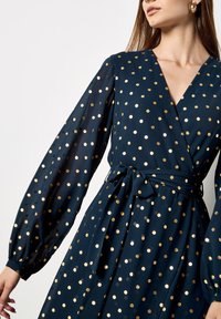 Abito avvolgente navy realizzato in tessuto morbido, con pois dorati, maniche lunghe a sbuffo e una cintura in vita per definire la silhouette.