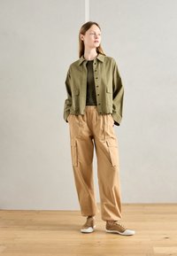 Olivgrüne, taillierte Jacke mit Knöpfen, beige Cargo-Hosen mit Taschen und hellbraune Sneakers mit weißer Sohle, auf einem Holzboden.