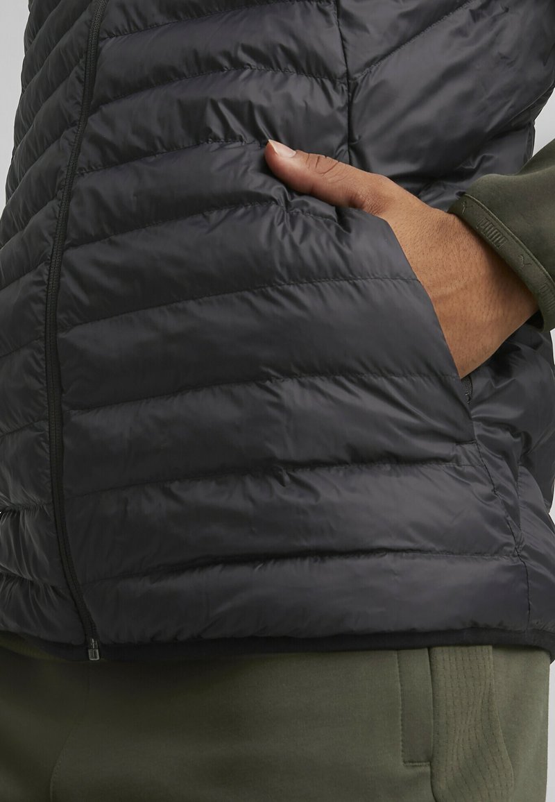 Doudoune noire sans manches avec couture matelassée, en matériau léger, fermeture éclair sur le côté et poche avant. Associée à un pantalon vert olive.