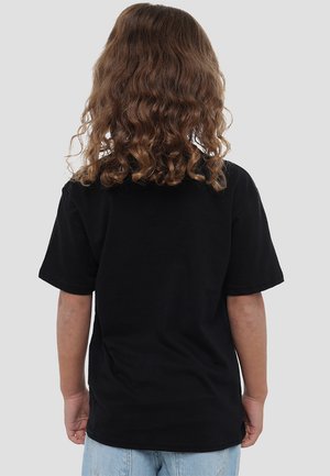 Ein Kind mit lockigem braunem Haar steht mit dem Rücken zur Kamera, trägt ein einfaches schwarzes T-Shirt und hellblaue Jeans vor einem grauen Hintergrund.