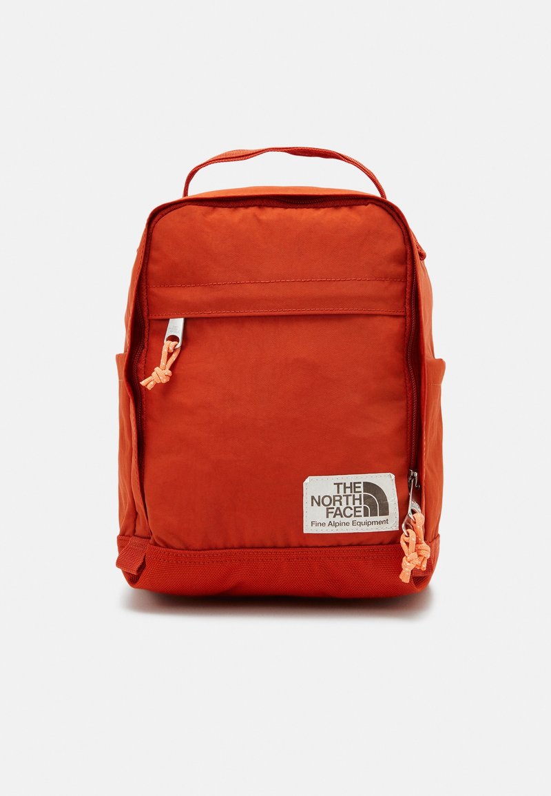 Sac à dos compact de forme carrée rouge avec poche zippée à l’avant, poignée supérieure, poches latérales et logo The North Face en bas à droite à l’avant.