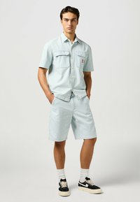 Lichtblauw gestreept kortweg shirt met twee borstzakken, gecombineerd met bijpassende shorts. Het model draagt zwart-witte sneakers met witte sokken.