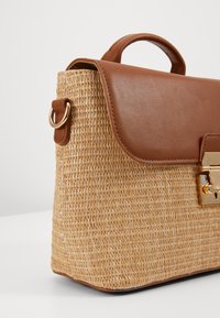 Borsa in paglia intrecciata con patta e manico in pelle marrone scuro, dotata di accessori in tono oro e dalla forma rettangolare. Testurizzata e resistente.