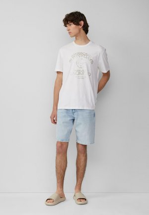 Weißes Baumwoll-T-Shirt mit Grafikdruck, hellblaue Jeansshorts und beiges Schuhwerk. Einfaches, lässiges Outfit mit weichen Texturen.