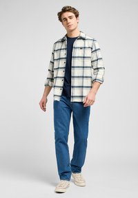 Camisa a cuadros en crema y azul marino, abotonada con un bolsillo en el pecho. Camiseta básica azul marino debajo. Vaqueros de mezclilla azul claro y zapatillas color beige.