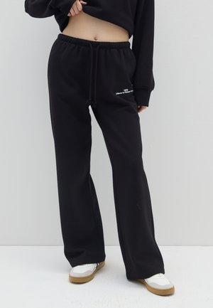 Pantaloni della tuta neri con una vestibilità ampia, cintura elastica e cordino. Testo bianco sulla parte frontale. Abbinati a sneakers bianche e un top nero.