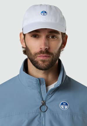 Homme barbu portant une casquette blanche et une veste à fermeture éclair bleu clair, toutes deux ornées de patches ronds avec le logo North Sails.