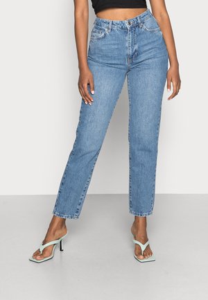 Kvinde iført højtaljede, blå jeans med lige ben, kombineret med lysegrønne sandaler med hæl, stående med det ene ben let bøjet.