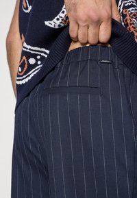 Hátulról látható tengerészkék csíkos nadrág, amelyen jól kivehető egy zseb és egy márkajelzés. Az anyag sima és jól strukturált hatást kelt.