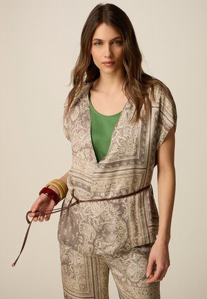 Kvinde iført beige bluse og bukser med paisley-mønster og en grøn top, der holder et brunt bælte og bærer tre store armbånd på sit håndled.