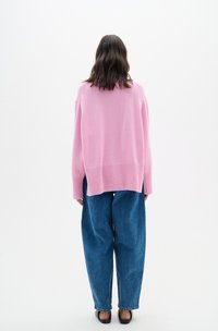 Maglione lavorato a maglia oversized di un rosa chiaro con orlo a costine, abbinato a pantaloni in denim blu larghi, visto da dietro su uno sfondo neutro.
