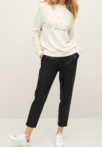 Kremowa bluza z napisem "C’est la vie" i "Paris 1990" w czerni, zestawiona z czarnymi spodniami i białymi trampkami. Gładka faktura materiału.