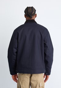 Veste bleu marine avec une texture lisse, col en velours côtelé et coutures visibles, accompagnée de pantalons cargo couleur beige.