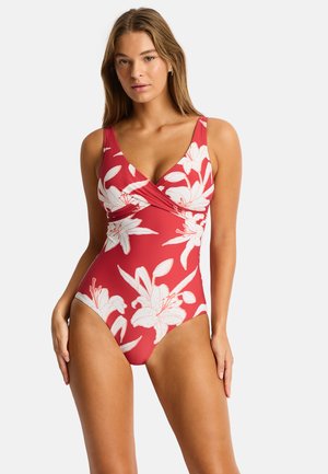 Femme portant un maillot de bain rouge une pièce avec un imprimé floral de lys blanc, debout, les bras relâchés sur un fond clair uni.