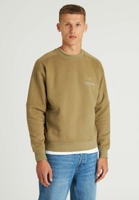 Felpa verde oliva con collo rotondo e polsini a coste, caratterizzata da un sottile dettaglio stampato sul petto. Abbinata a jeans azzurri chiari.