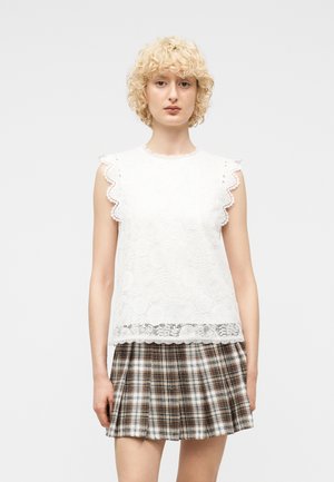 Joven con cabello rizado y rubio corto que lleva una blusa blanca sin mangas de encaje y falda plisada de cuadros marrones, de pie frente a un fondo liso.