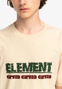 Beige bomuld t-shirt med fed grøn og maroon tekst, med et grafisk design der integrerer bladmønstre i bogstaverne.