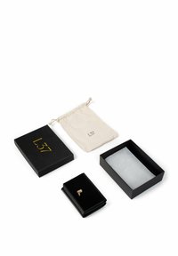 Portafoglio in pelle nera con dettaglio dorato, posizionato accanto a una scatola nera con scritte dorate, una bustina bianca con coulisse e un panno grigio.