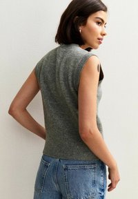 Jonge vrouw met kort donker haar, gekleed in een mouwloze grijze gebreide top en blauwe denim jeans, die zijwaarts poseert tegen een witte muur.