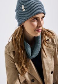 Berretto a costine blu con un piccolo etichetta, abbinato a una sciarpa tubolare blu coordinata, indossato sopra una maglietta nera sotto un cappotto beige.