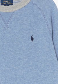 Ljusblå sweatshirt med grå krage, med en liten svart polo-spelare logga på bröstet. Materialet verkar mjukt och strukturerat.