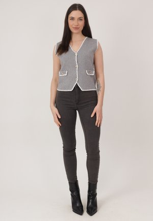 Kobieta ubrana w szarą, bezrękawnikową kamizelkę zapinaną na guziki z białym obszyciem, czarne obcisłe dżinsy oraz czarne botki, stojąca na tle jednolitego tła.