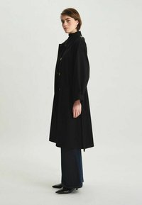Long manteau noir à coupe décontractée, avec de grands boutons, des poches latérales et des poignets retroussés, porté sur un jean foncé et des chaussures noires à bout pointu.