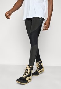 Μαύρο και σκούρο γκρι αθλητικό κολάν με εφαρμοστή σχεδίαση, που διαθέτει αντίθετο πάνελ και λογότυπο κοντά στη μέση, σε συνδυασμό με μαύρα και καφέ αθλητικά παπούτσια.