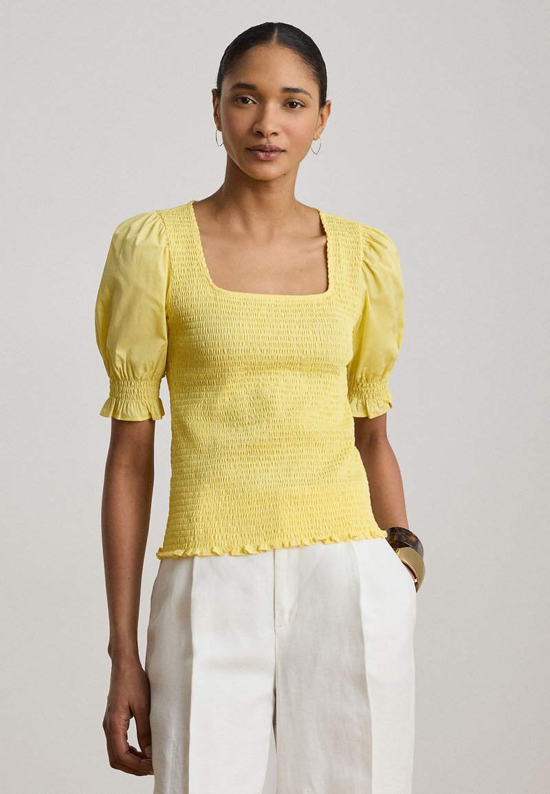 Γυναίκα που φοράει κίτρινο τοπ με σφηκοφωλιά, τετράγωνη λαιμόκοψη και φουσκωτά μανίκια, καθώς και λευκό παντελόνι με ψηλή μέση, στέκεται μπροστά από απλό ανοιχτό φόντο.