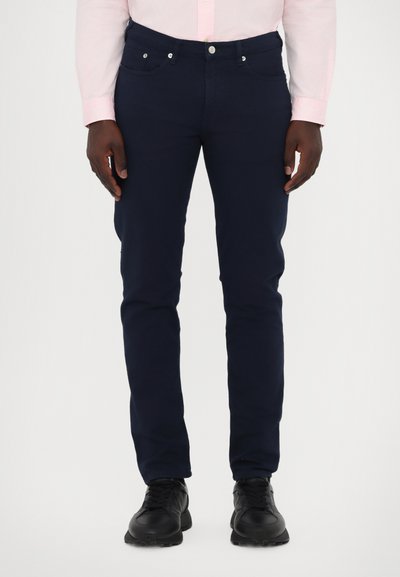 Jeans en coton bleu marine avec une coupe slim, comportant cinq poches, un bouton en métal et une fermeture éclair. Des chaussures de sport noires complètent le look.