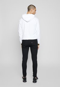 Pessoa com cabelo muito curto vestindo um moletom branco, jeans skinny pretos e ténis pretos, de pé, de costas, contra um fundo simples.