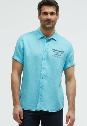 Mann trägt ein hellblaues kurzärmeliges Hemd mit aufgekrempelten Ärmeln und marineblaue Hose vor einem einfarbigen Hintergrund.